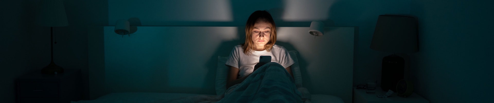 Sleepy exhausted woman lying in bed using smartphone, can not sleep. Insomnia, addiction concept. Sad girl bored in bed scrolling through social networks on mobile phone late at night in dark bedroom.
